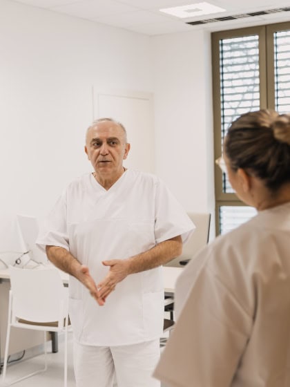 A doctor in a white medical coat is talking to a person in a beige uniform in a modern hospital room.<br>