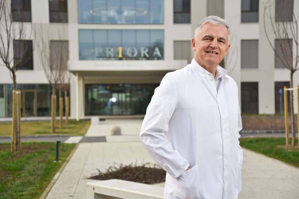 Stariji muškarac u bijeloj liječničkoj kuti stoji ispred moderne zgrade s natpisom 'PRIORA' iznad ulaza.