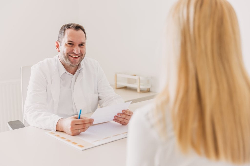 A doctor in a white coat sits at a desk, holding papers and speaking with a female patient whose blonde hair is visible from behind.
