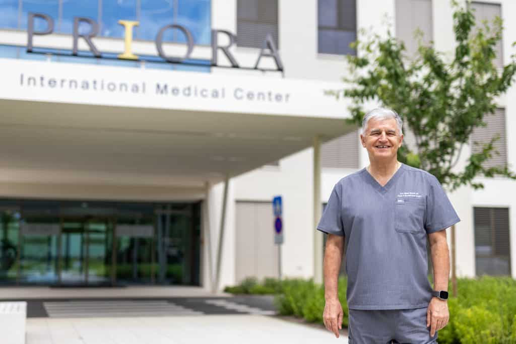 A man in medical clothing stands in front of a building with the sign 'International Medical Center'.