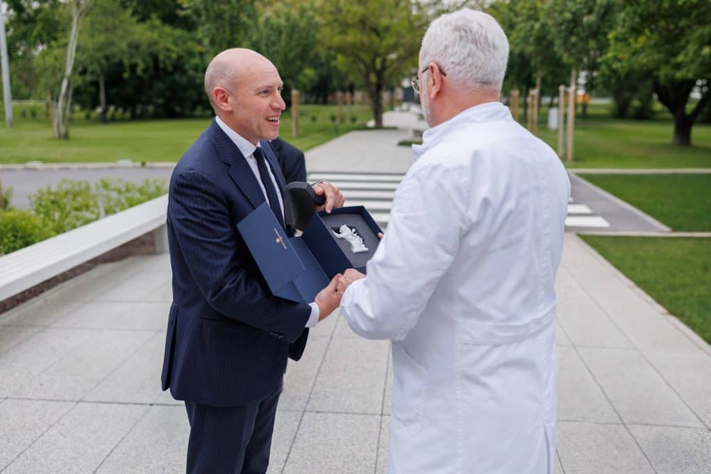 Muškarac u odijelu prima nagradu u plavoj kutiji od muškarca u bijelom laboratorijskom ogrtaču na otvorenom, na pločniku okruženom zelenilom.