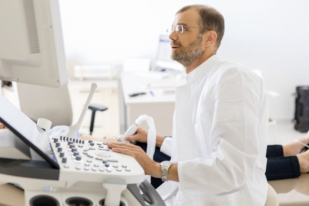 A man in a white medical coat performs an ultrasound using a machine in a bright examination room.