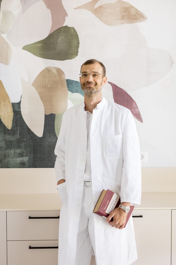 A man in a white coat, holding a book, stands next to a colorful wall design.