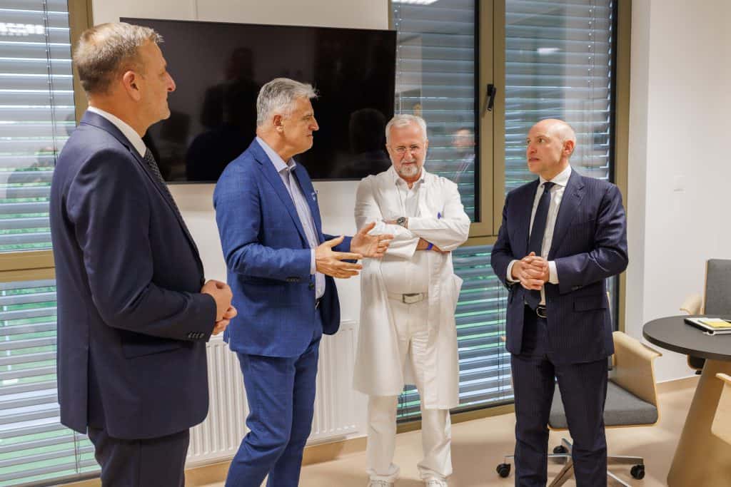 Četiri muškarca u poslovnim i liječničkim odijelima stoje u sobi i razgovaraju; iza njih je zid s prozorima i ravnim televizorom.