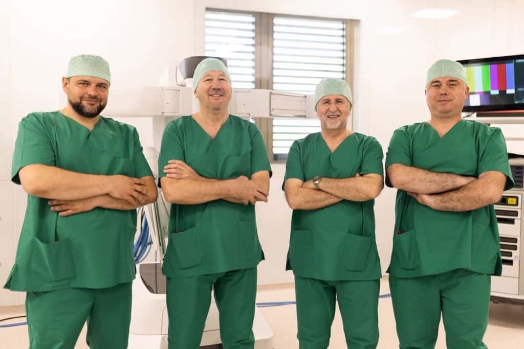 Četiri muškarca u zelenim kirurškim odorama stoje s prekriženim rukama u operacijskoj sali.
