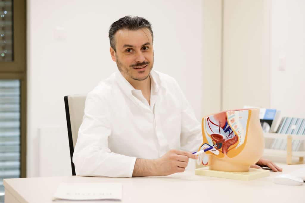 A man in a white shirt sits at a desk, pointing to a medical model of the pelvis in an office setting.