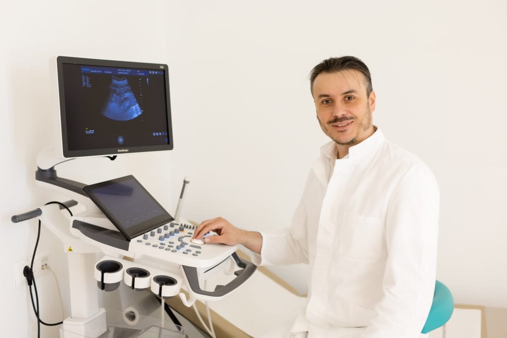 A man in white medical attire sits next to an ultrasound machine, looking at the camera and operating the device’s control panel.