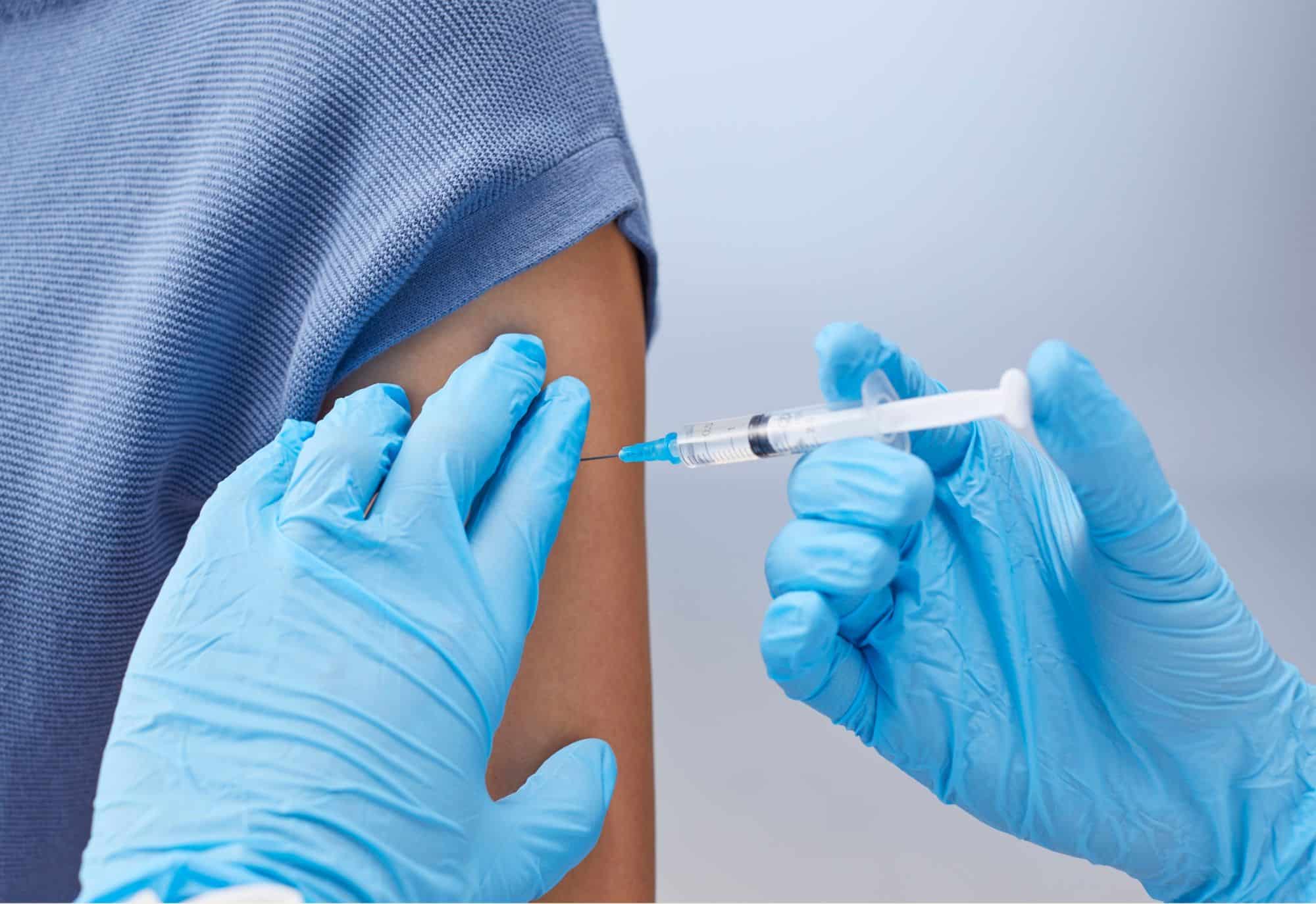Gloved healthcare worker administers a vaccine injection into a person’s upper arm with their sleeve rolled up.