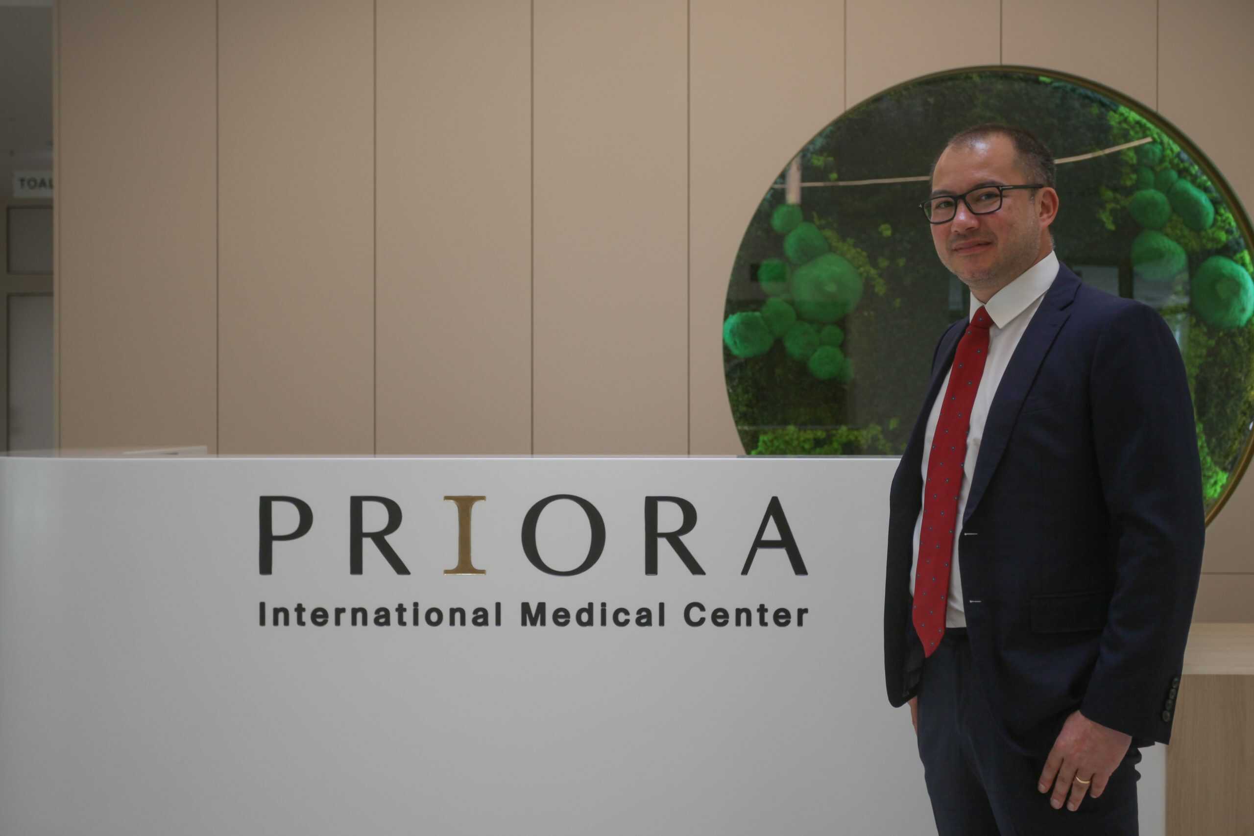 A man in a dark suit with a red tie stands by the reception desk with the sign “PRIORA International Medical Center,” in front of a circular green wall covered with plants.<br>