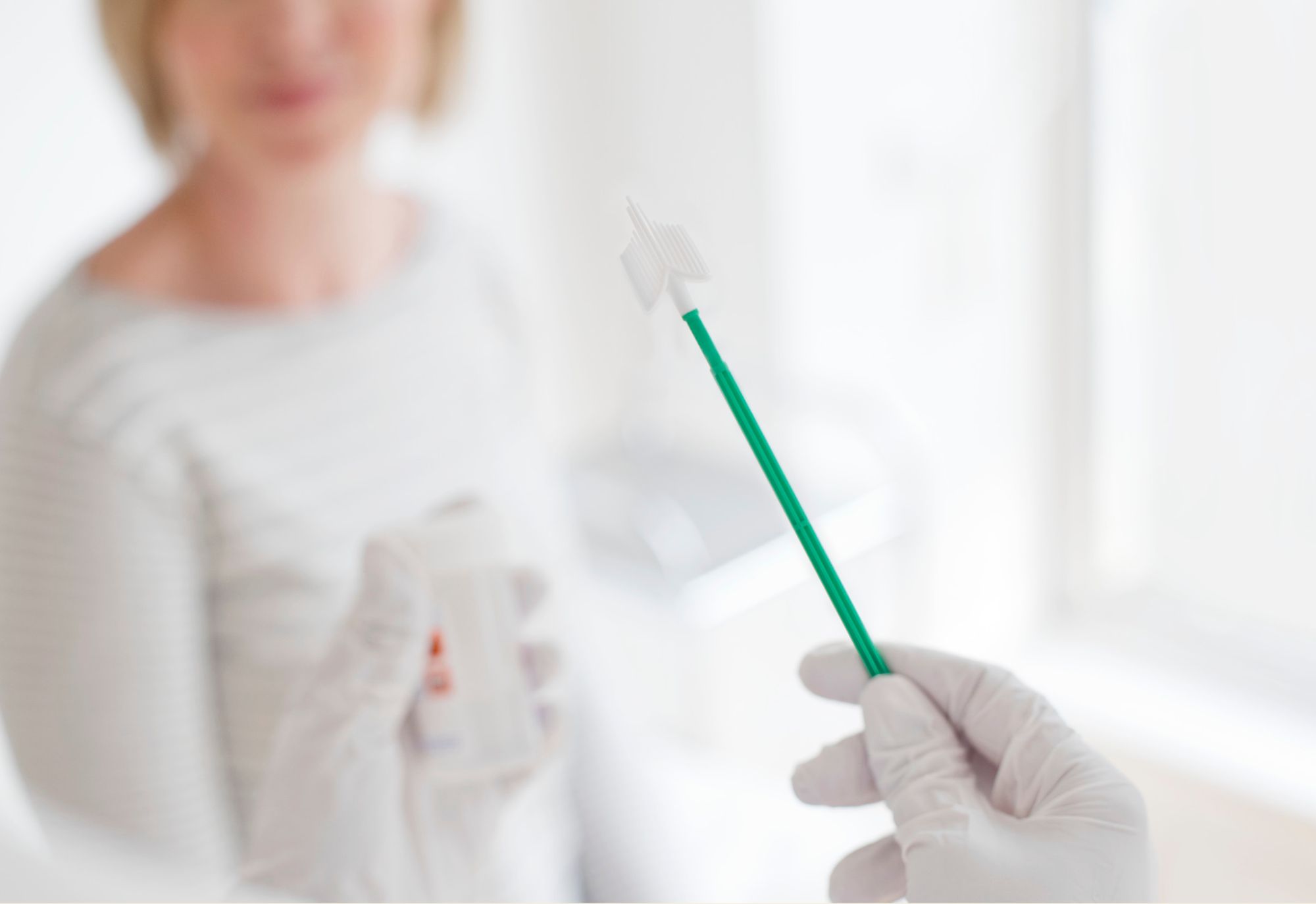 Gloved hand holding a cervical brush for collecting a sample (Pap smear), with a patient and sample vial blurred in the background.