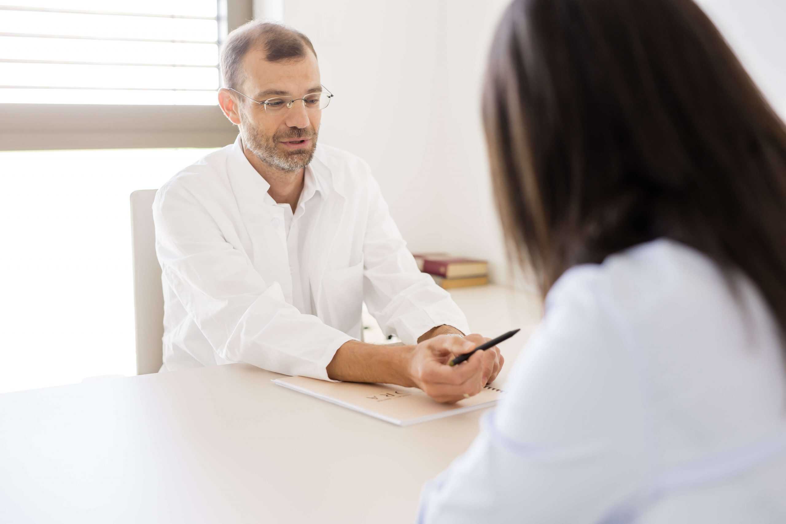 Liječnik u bijeloj kuti za stolom objašnjava nešto pacijentu, držeći olovku iznad papira u svijetloj ordinaciji.
