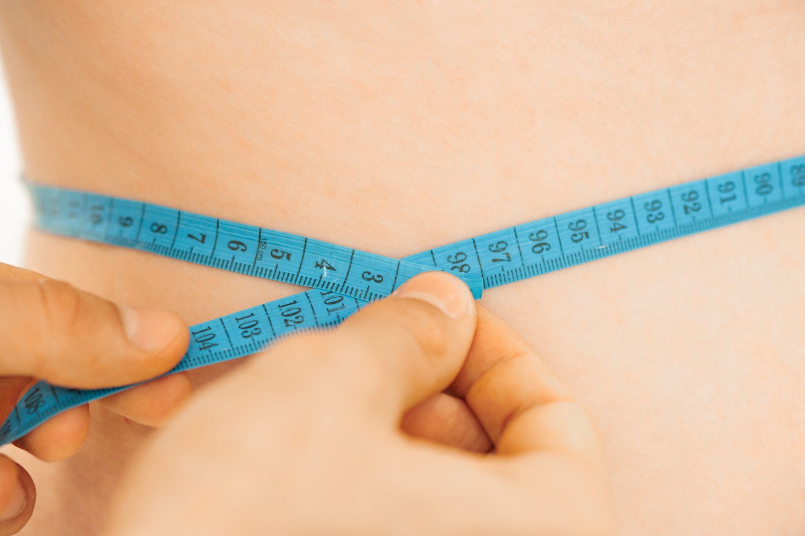 A close-up of hands tightening a blue measuring tape around a person’s waist, measuring circumference.