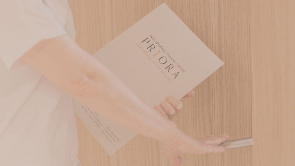 A person holds a brochure with the words "PRIORA - International Medical Center" as he opens the door.