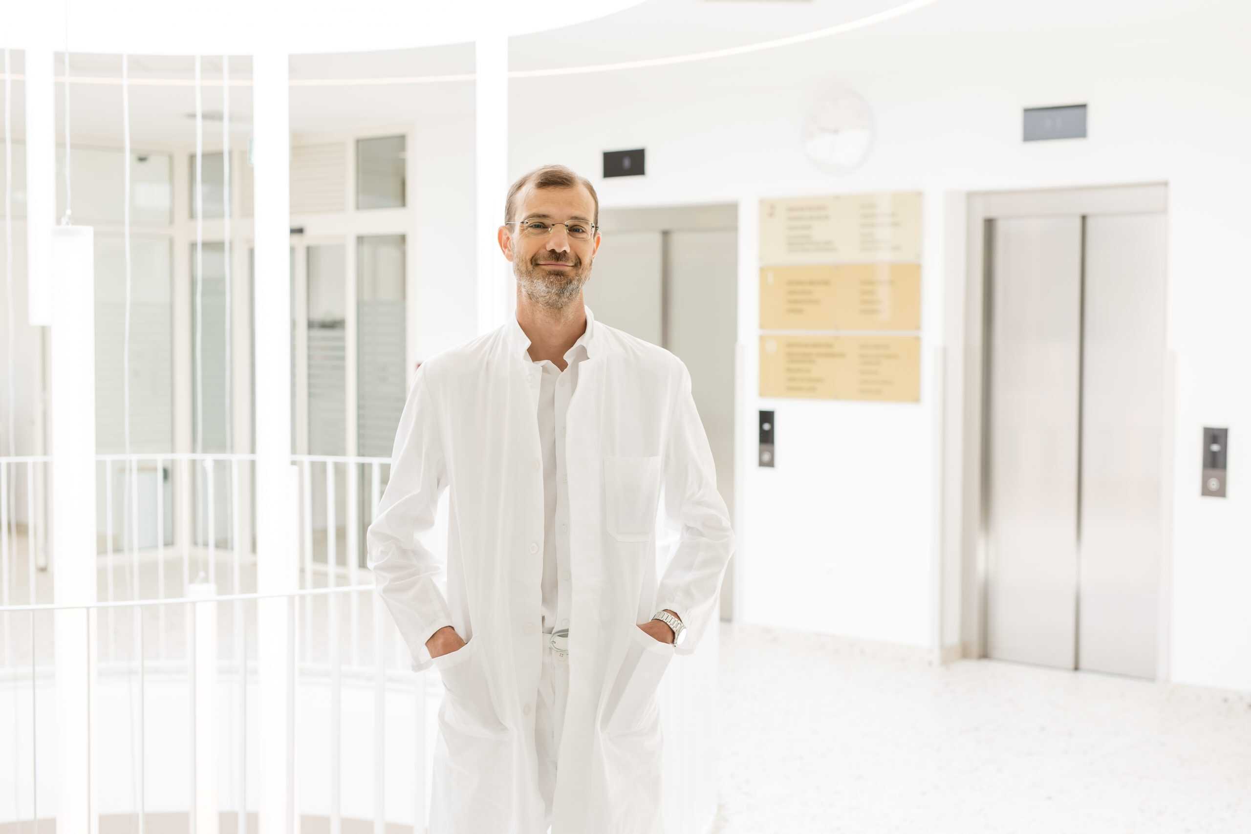 A man in a white medical coat, wearing glasses and a short beard, stands in a bright hospital hallway in front of an elevator, with his hands in his pockets.