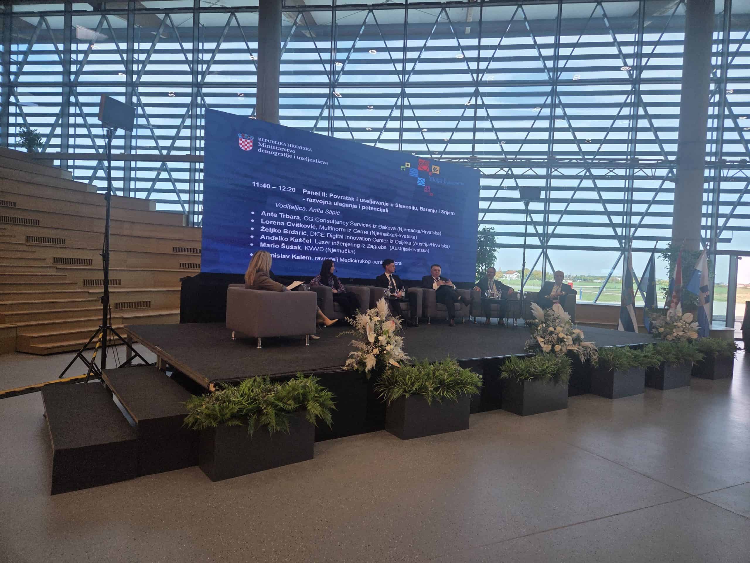 Panel-rasprava na pozornici s više sudionika u modernoj staklenoj dvorani, ispred velike plave projekcije s programom na hrvatskom, uz zastave i cvjetne aranžmane duž ruba pozornice.