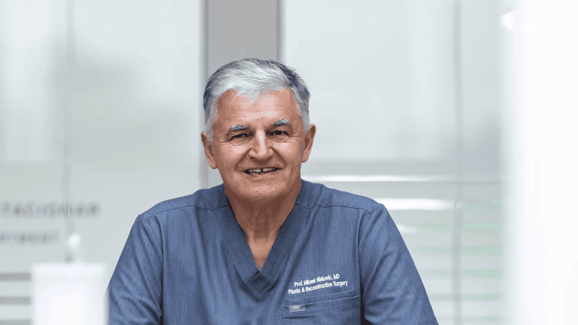 Portret starijeg muškarca sijede kose u plavoj medicinskoj uniformi, nasmiješen u svijetloj klinici, s natpisom „Plastic & Reconstructive Surgery” na džepu.