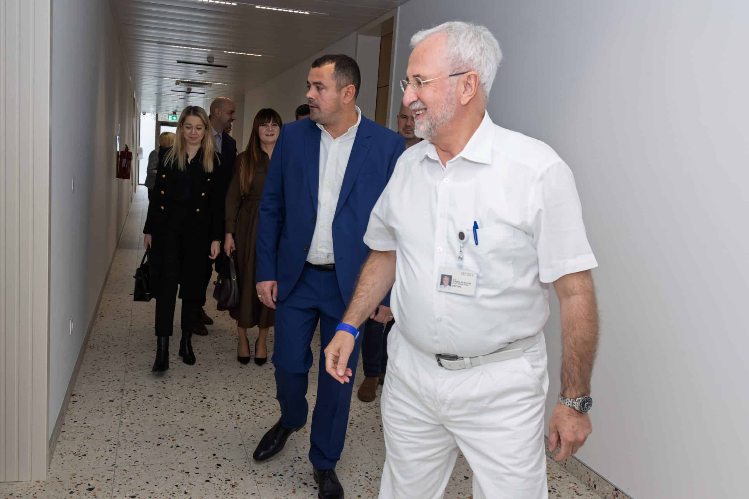 Liječnik u bijeloj uniformi s identifikacijskom iskaznicom vodi skupinu posjetitelja kroz svijetli bolnički hodnik, dok ga prate muškarac u plavom odijelu i nekoliko žena.