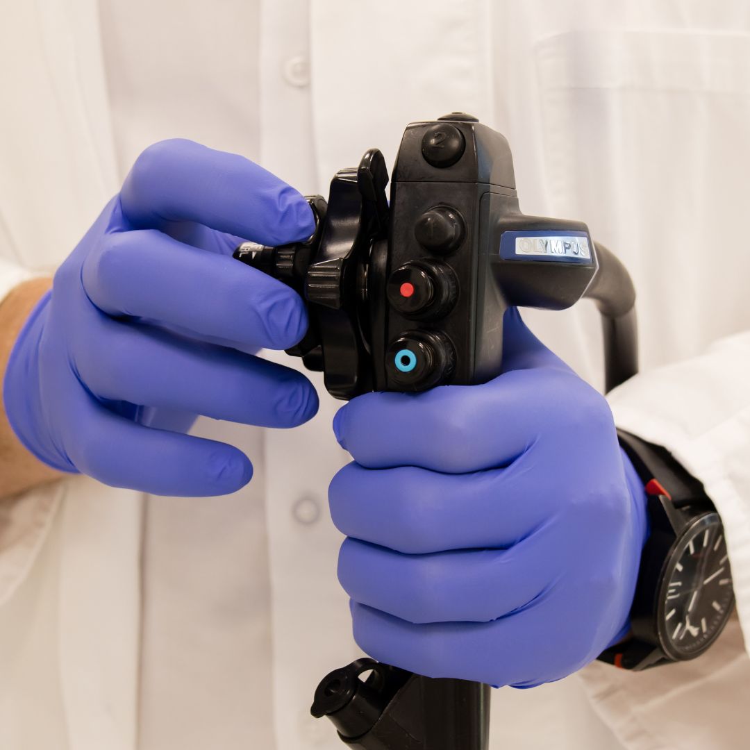 Ruke u plavim rukavicama drže kontrolnu ručku endoskopa s gumbima i kotačićima.