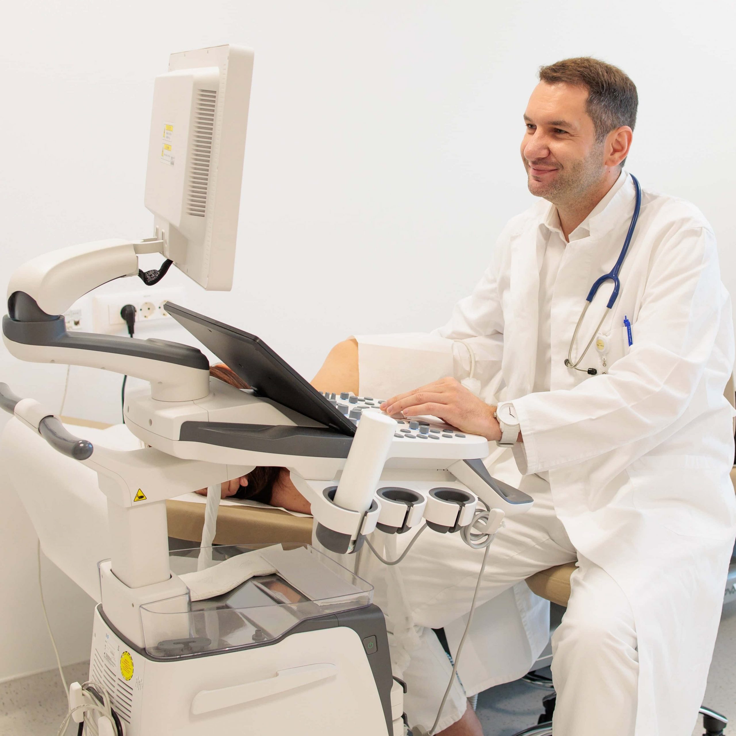 Liječnik u bijeloj kuti sa stetoskopom upravlja ultrazvučnim aparatom tijekom pregleda pacijenta u ordinaciji.