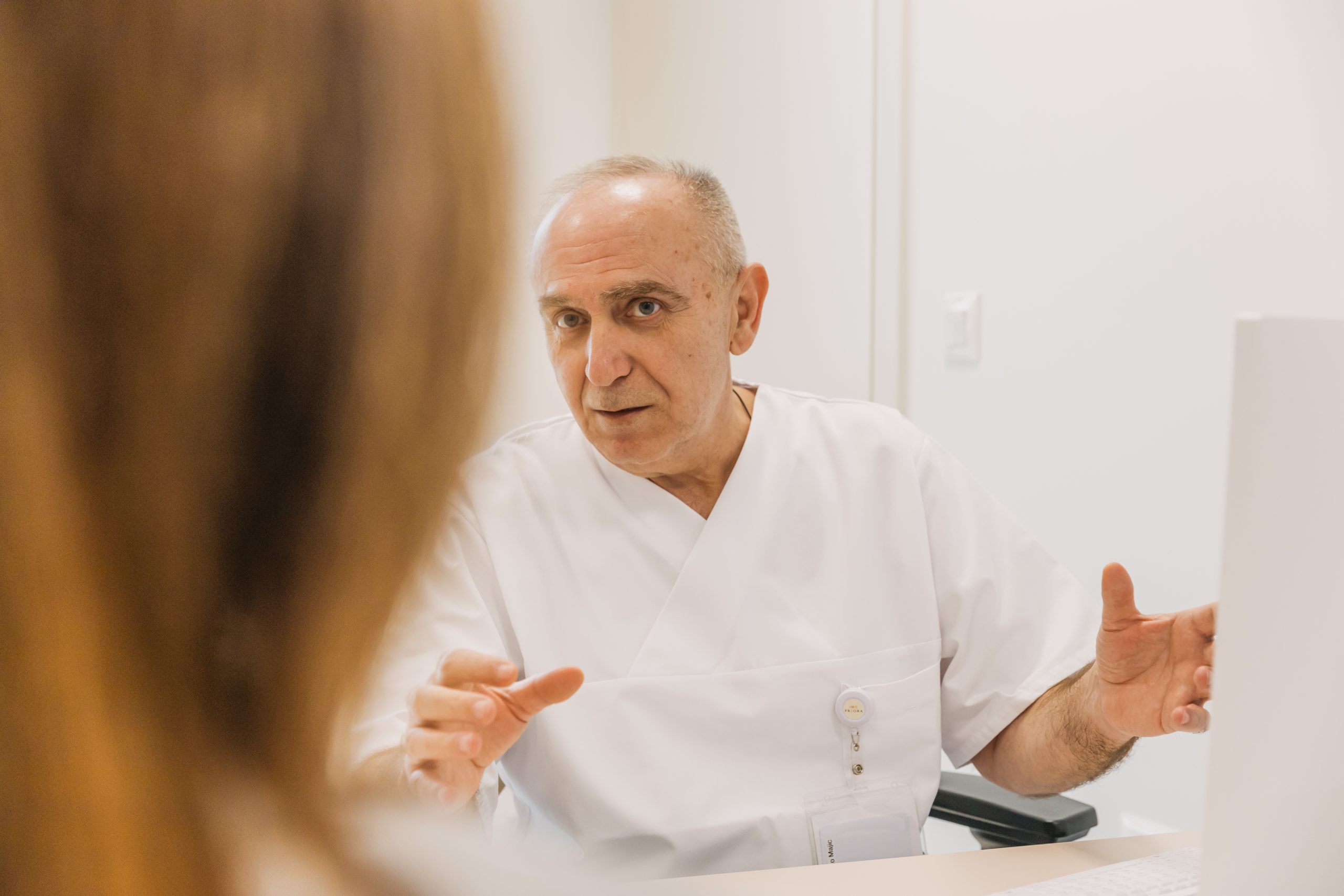 Liječnik u bijeloj uniformi gestikulira dok razgovara s pacijentom u ordinaciji, pacijent je izvan fokusa u prvom planu.