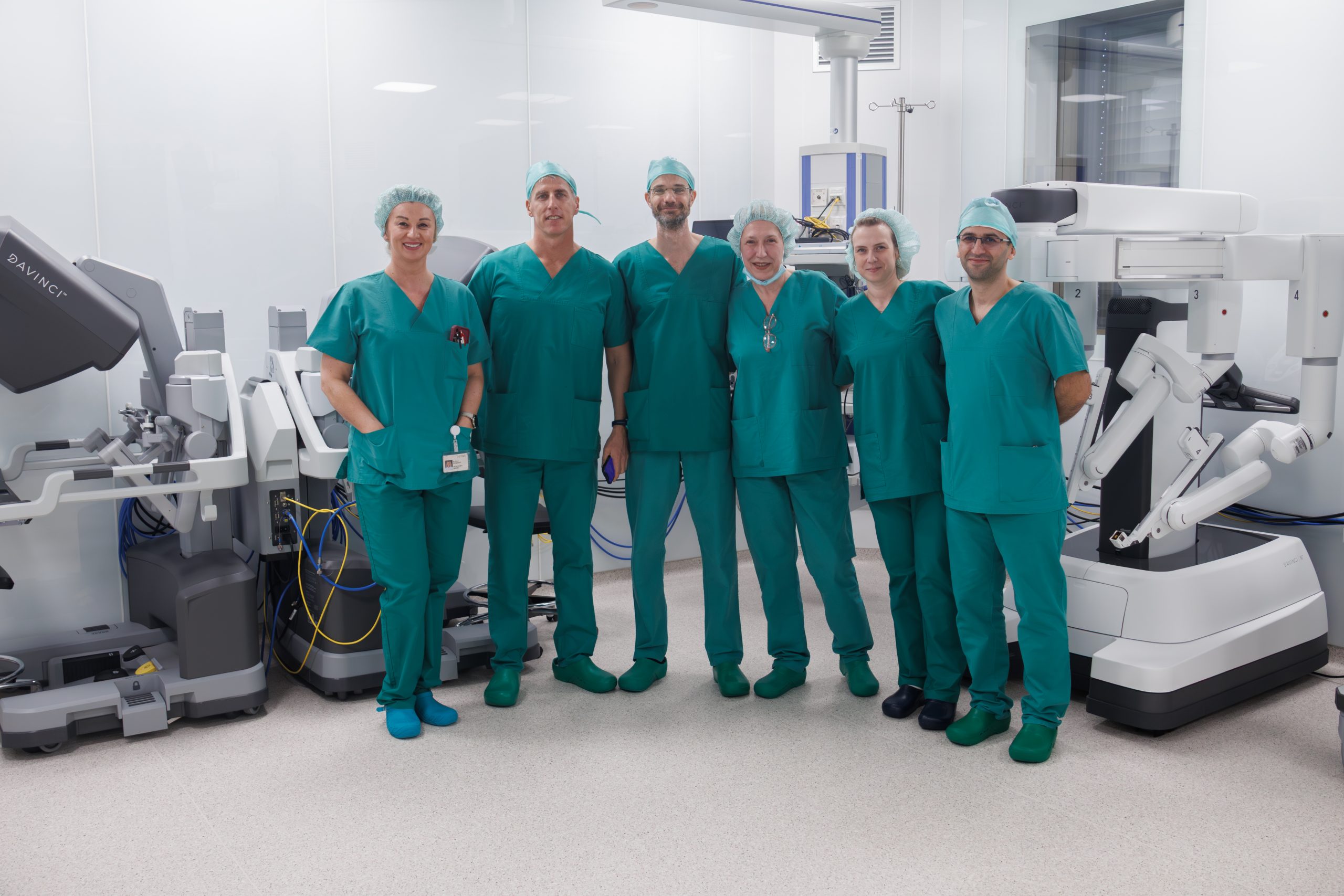 Šesteročlani kirurški tim u zelenim odorama stoji u operacijskoj sali s opremom za robotsku kirurgiju.