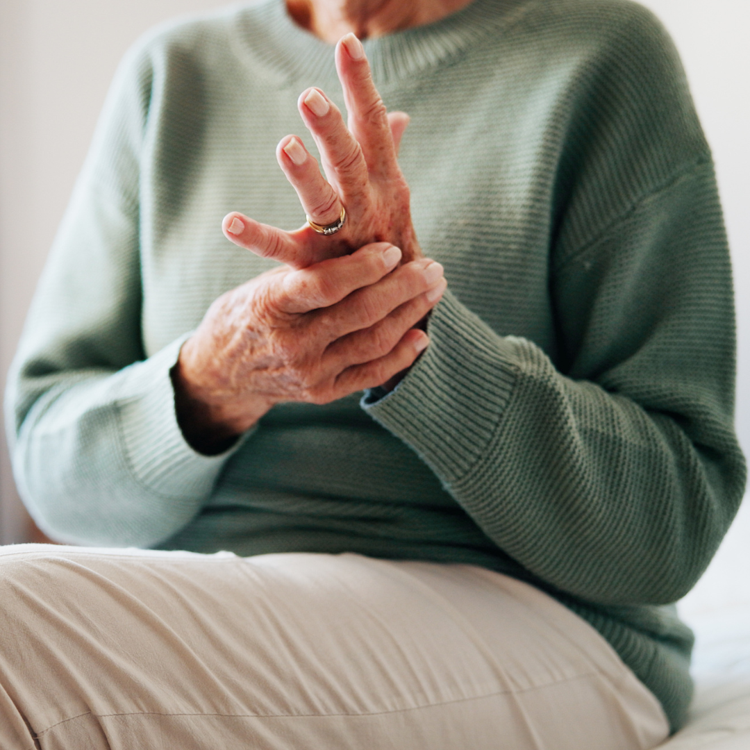 Osoba u zelenom puloveru sjedi i drži lijevu ruku desnom, fokus na naborane ruke s prstenom.