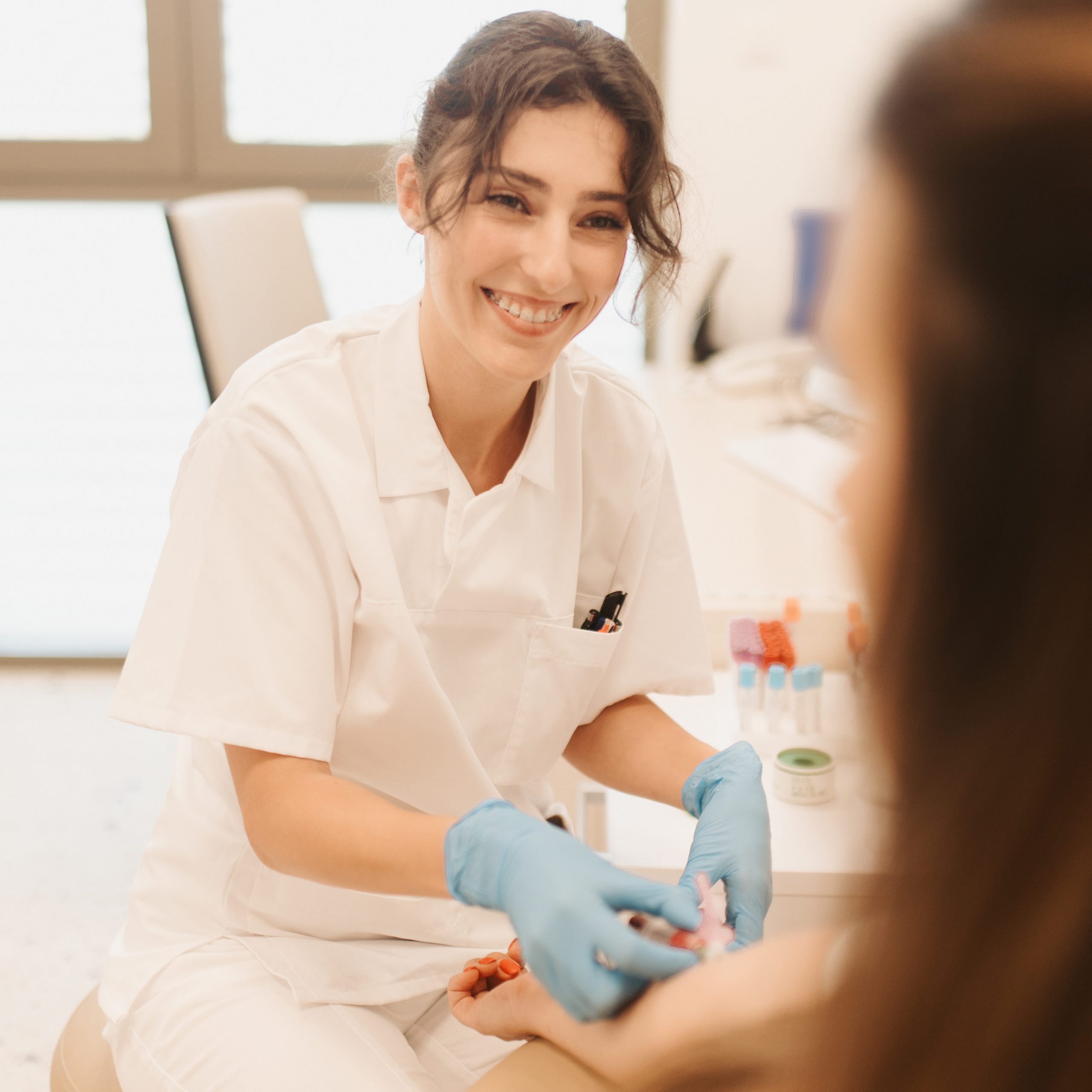 Zdravstveni djelatnik u bijeloj uniformi i plavim rukavicama uzima uzorak krvi pacijentu u klinici, srdačno se smiješi.