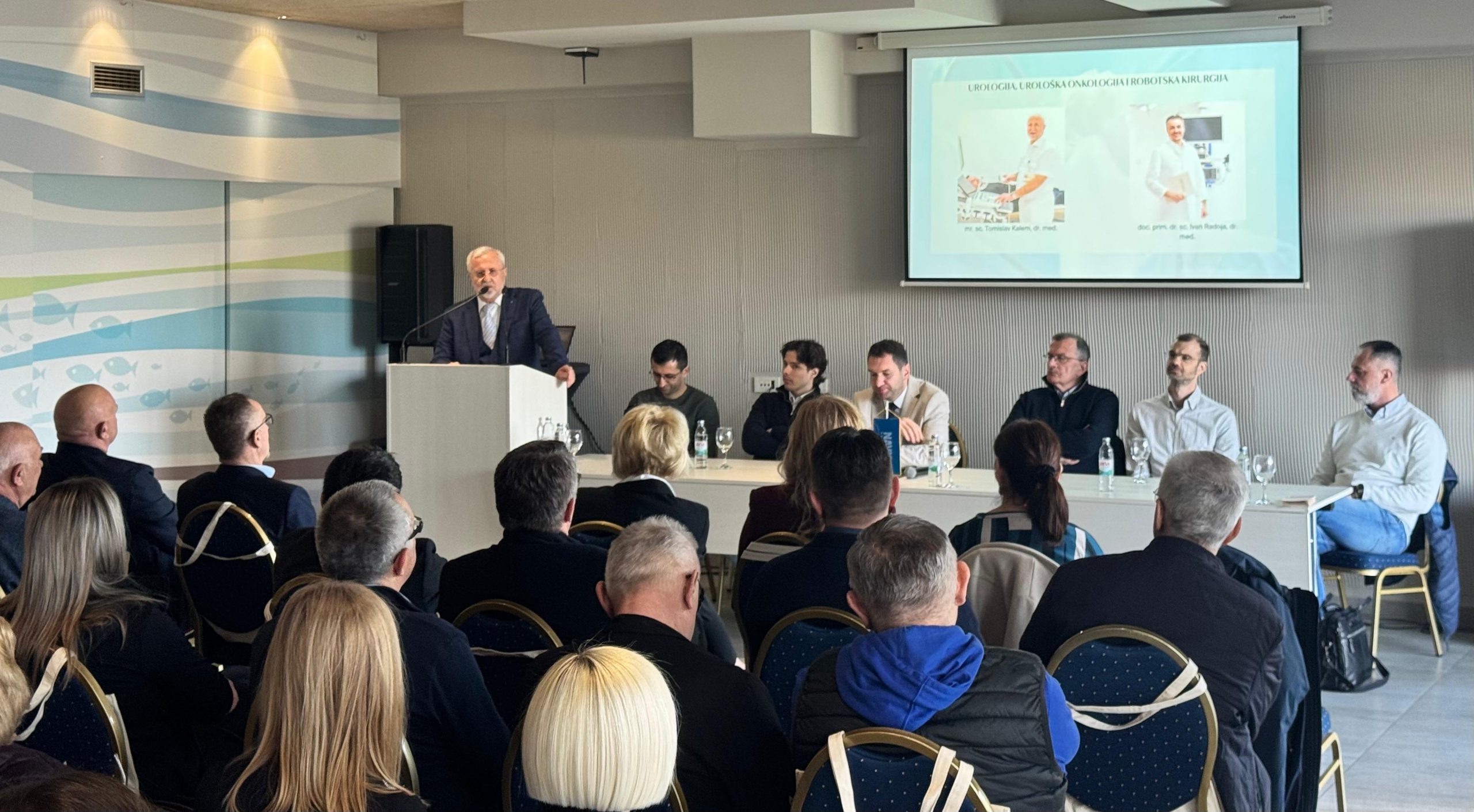 Govornik za pultom obraća se publici u konferencijskoj dvorani, uz panel stručnjaka za stolom i prezentaciju projiciranu na zidu.