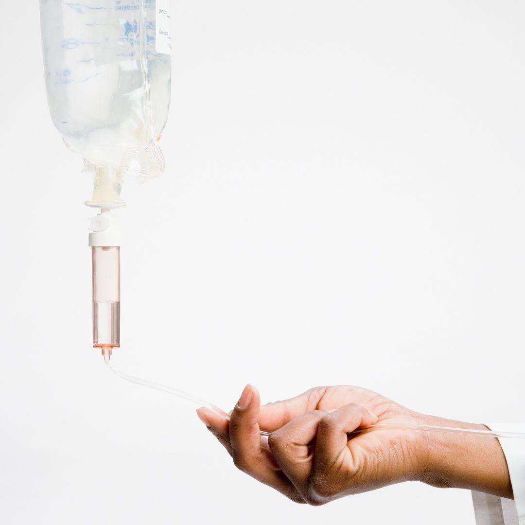 Ruka tamnije puti drži cjevčicu infuzije povezanu s visećom vrećicom na bijeloj pozadini.