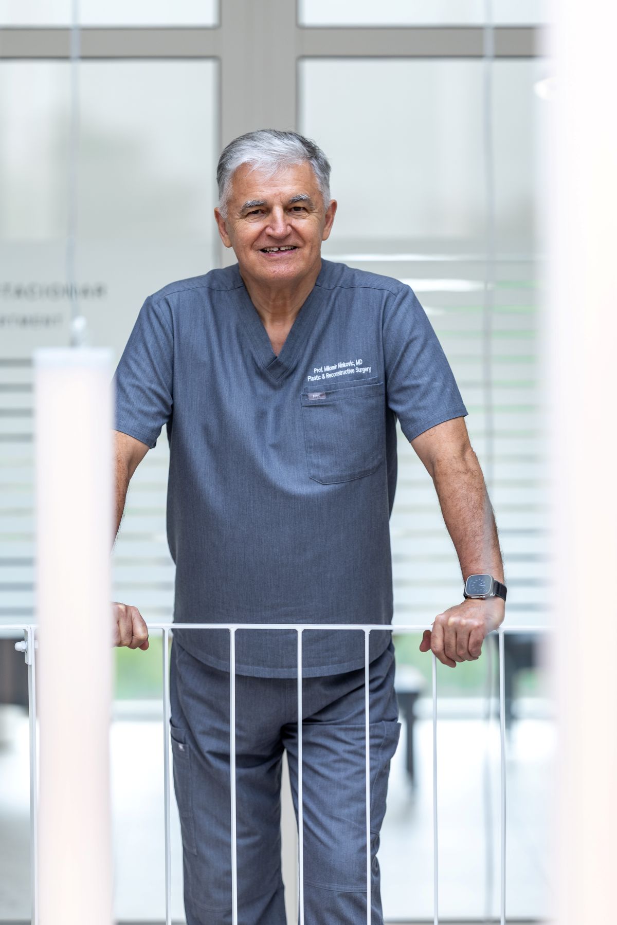 Stariji muškarac u sivom medicinskom odijelu nasmiješeno stoji u svijetloj klinici, držeći se za bijeli rukohvat.