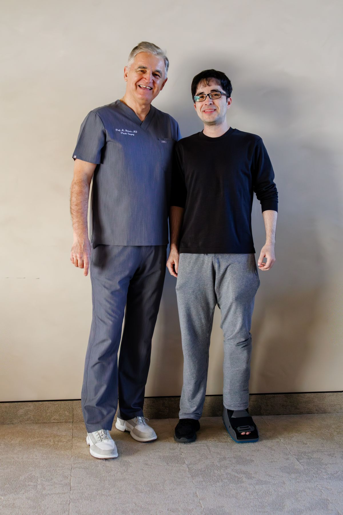 Dvojica muškaraca stoje nasmiješeni; lijevi u sivim medicinskim odorama, desni u crnoj majici i trenirci, s ortozom na lijevoj nozi.