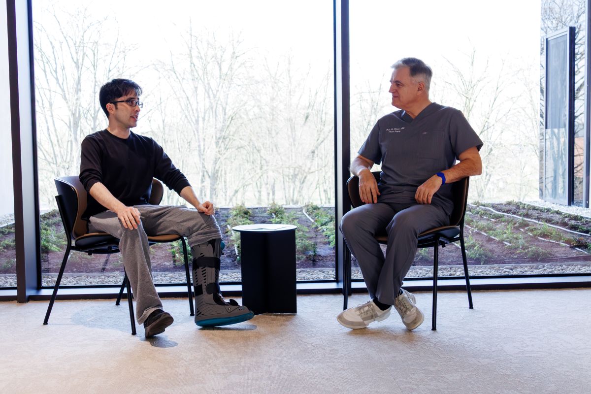 Dvoje ljudi sjedi i razgovara; lijevi muškarac nosi ortopedsku čizmu na nozi, desni u medicinskoj uniformi, pozadina s prozorom.