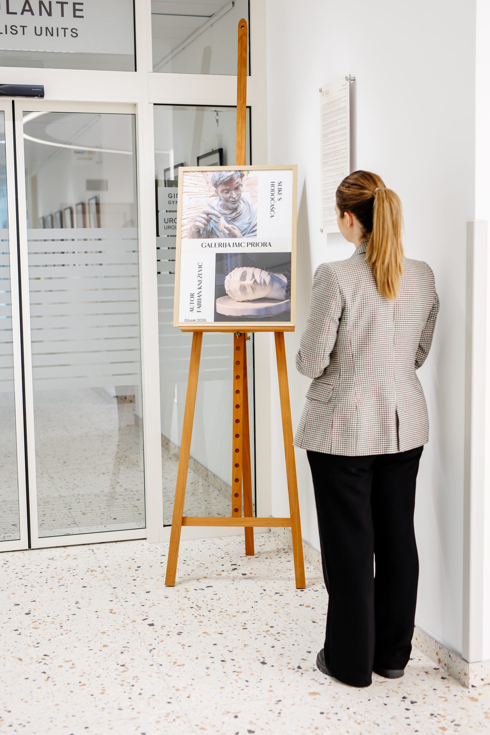 Žena s repom gleda plakat na stalku u galeriji; na plakatu su umjetnički portreti i tekst “GALERIJA DE PROUBA”.