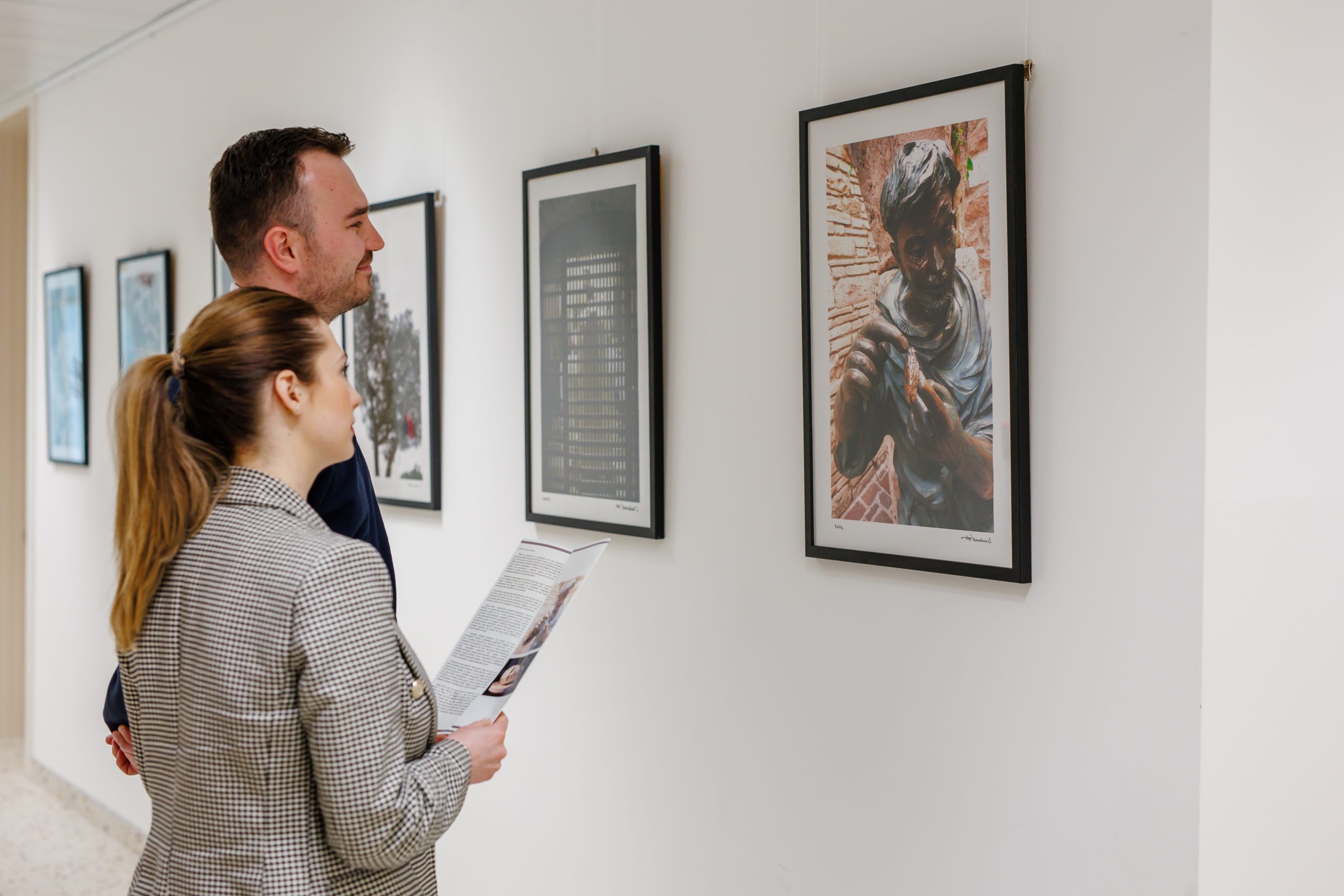 Dvoje ljudi u galeriji gledaju uokvirenu fotografiju skulpture, dok žena drži tiskani vodič izložbe.