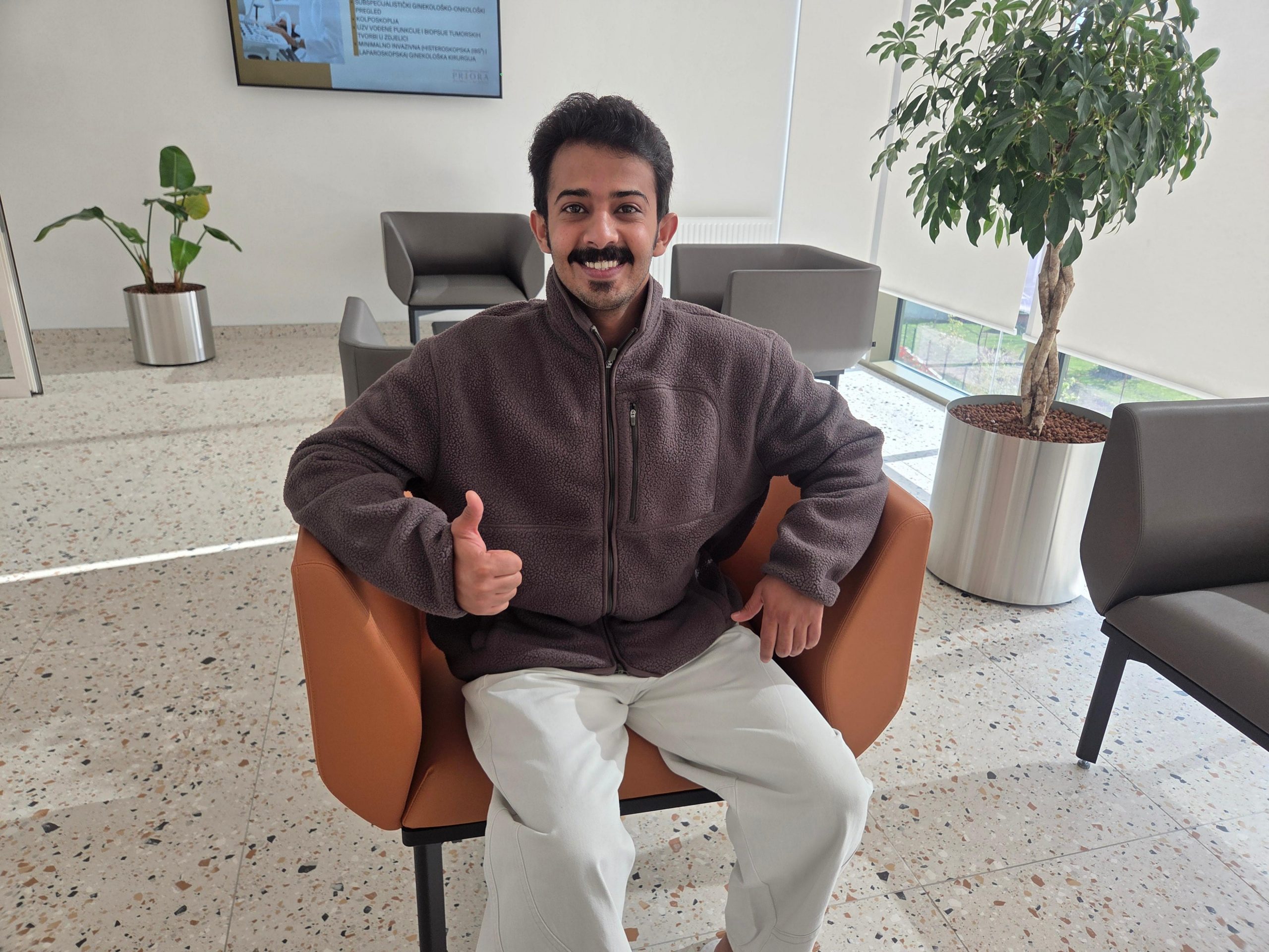 Muškarac sjedi u modernoj čekaonici, nasmijan i podiže palac gore, okružen biljkama u metalnim teglama i sivim foteljama.