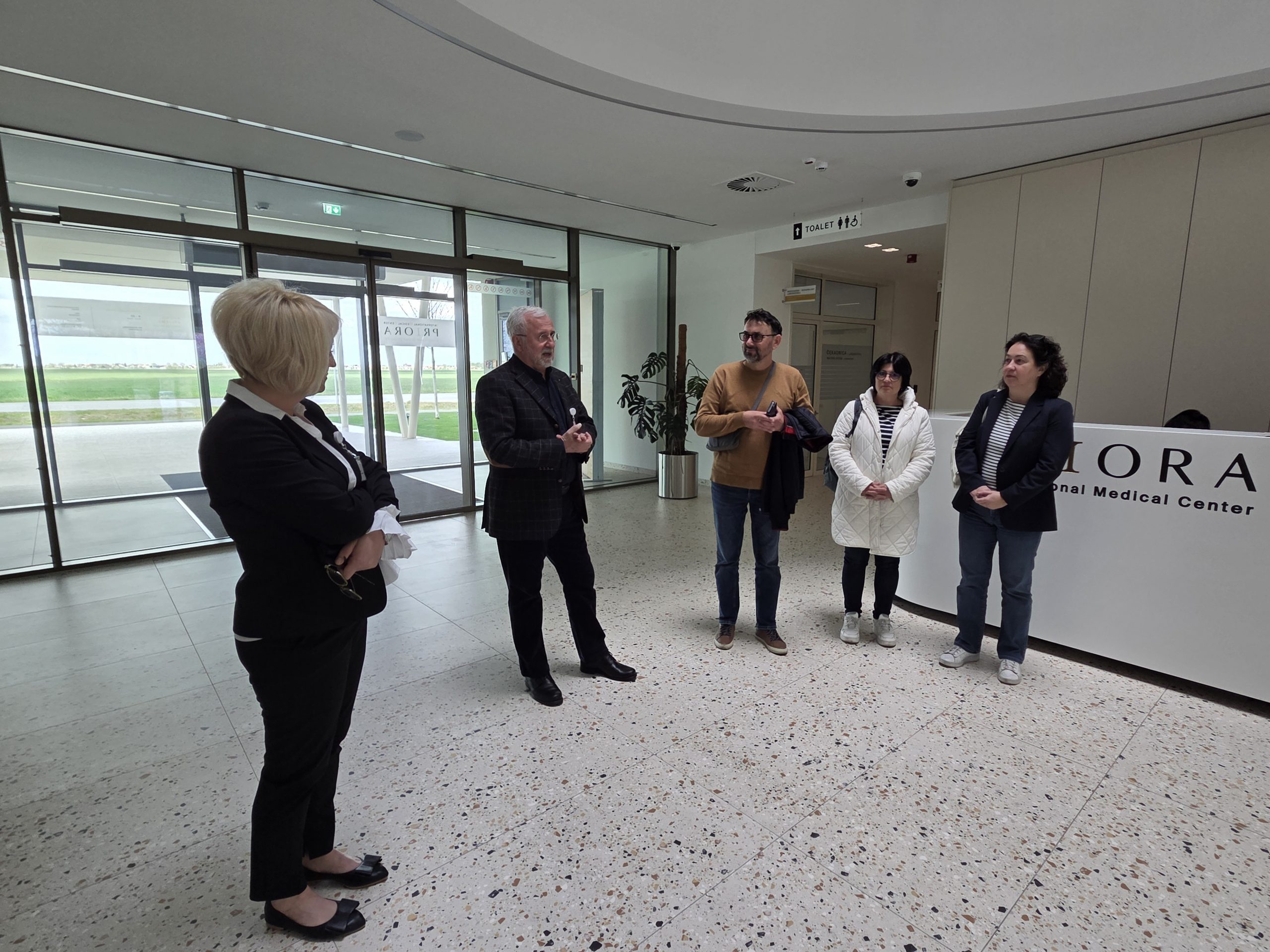 Skupina pet ljudi razgovara u modernom predvorju medicinskog centra, s recepcijskim pultom i staklenim zidom u pozadini.