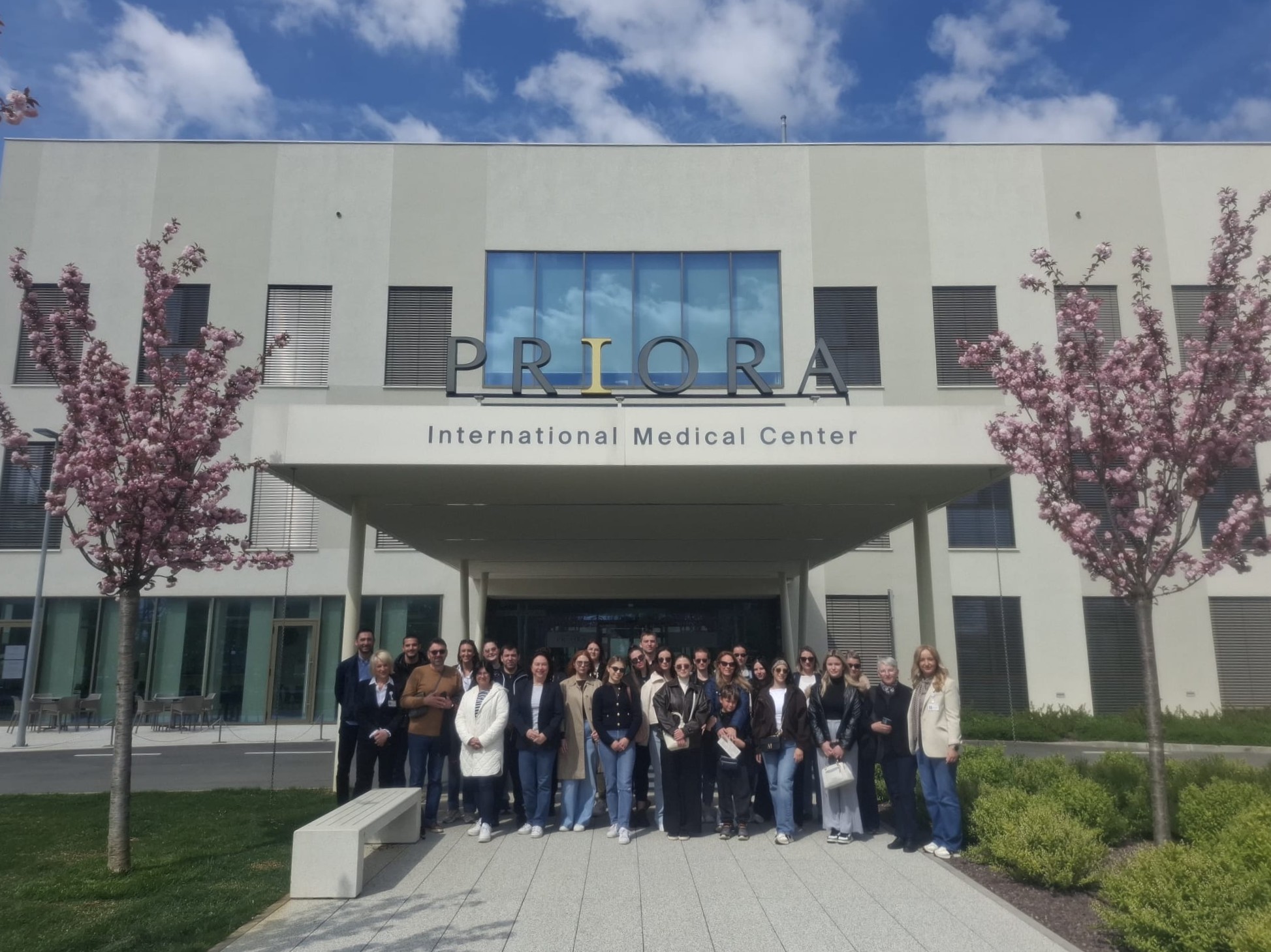 Skupina ljudi pozira ispred ulaza u Priora International Medical Center, s procvalim trešnjama i sunčanim nebom.