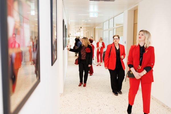 Skupina ljudi, mnogi u crvenoj odjeći, razgleda uokvirene radove u svijetlom hodniku.