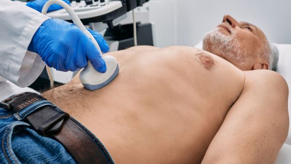 Stariji muškarac leži dok medicinski radnik u plavim rukavicama radi ultrazvuk abdomena ručnom sondom.