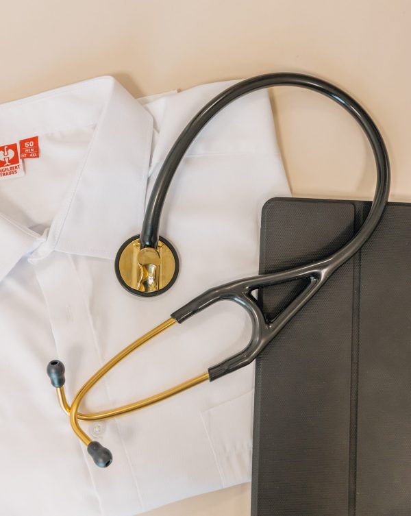 Crni stetoskop sa zlatnim dijelovima položen na bijeloj košulji pokraj crne futrole za tablet.