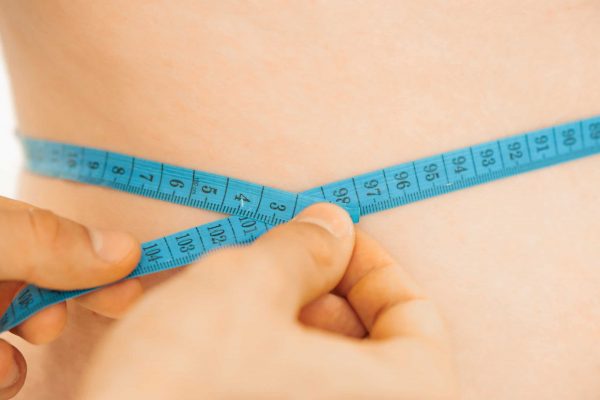 Krupni plan ruku koje zatežu plavu metarsku traku oko struka osobe, mjereći opseg.