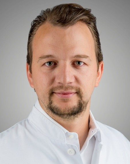 Portret muškarca u bijeloj liječničkoj kuti, neutralna pozadina, blag osmijeh i kratka brada.