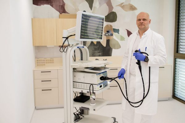 Liječnik u bijeloj kuti i plavim rukavicama drži endoskop pokraj medicinskog kolonoskopskog sustava u svijetloj ordinaciji.