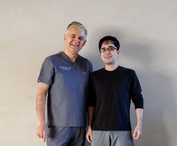 Dvojica muškaraca stoje rame uz rame; stariji u sivom medicinskom odijelu s osmijehom, mlađi u crnoj majici, neutralna pozadina.