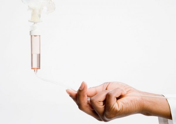 Ruka koja drži cijev infuzije s prozirnom komorom ispunjenom blijedoružičastom tekućinom na bijeloj pozadini.