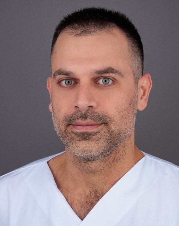 Portret muškarca kratke tamne kose i brade u bijeloj medicinskoj uniformi, neutralna siva pozadina.