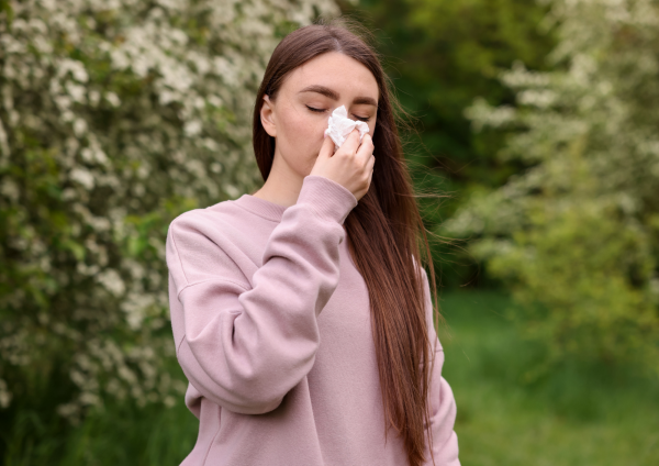 Mlada žena na otvorenom briše nos maramicom, zatvorenih očiju, okružena cvatućim grmljem i zelenilom.