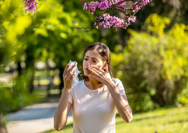 Žena na otvorenom kiše i briše nos maramicom ispod cvatućeg stabla, okružena zelenilom u proljeće.