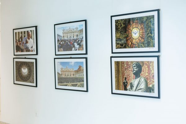 Bijeli galerijski zid s dvoredno izloženim uokvirenim fotografijama Vatikana: bazilika, mnoštvo vjernika, vitraji i kip s aureolom.