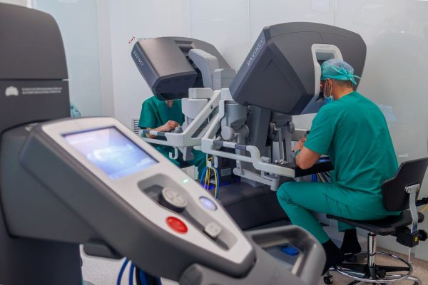 Two surgeons in green scrubs operate a robotic surgery console in a modern, sterile medical room.